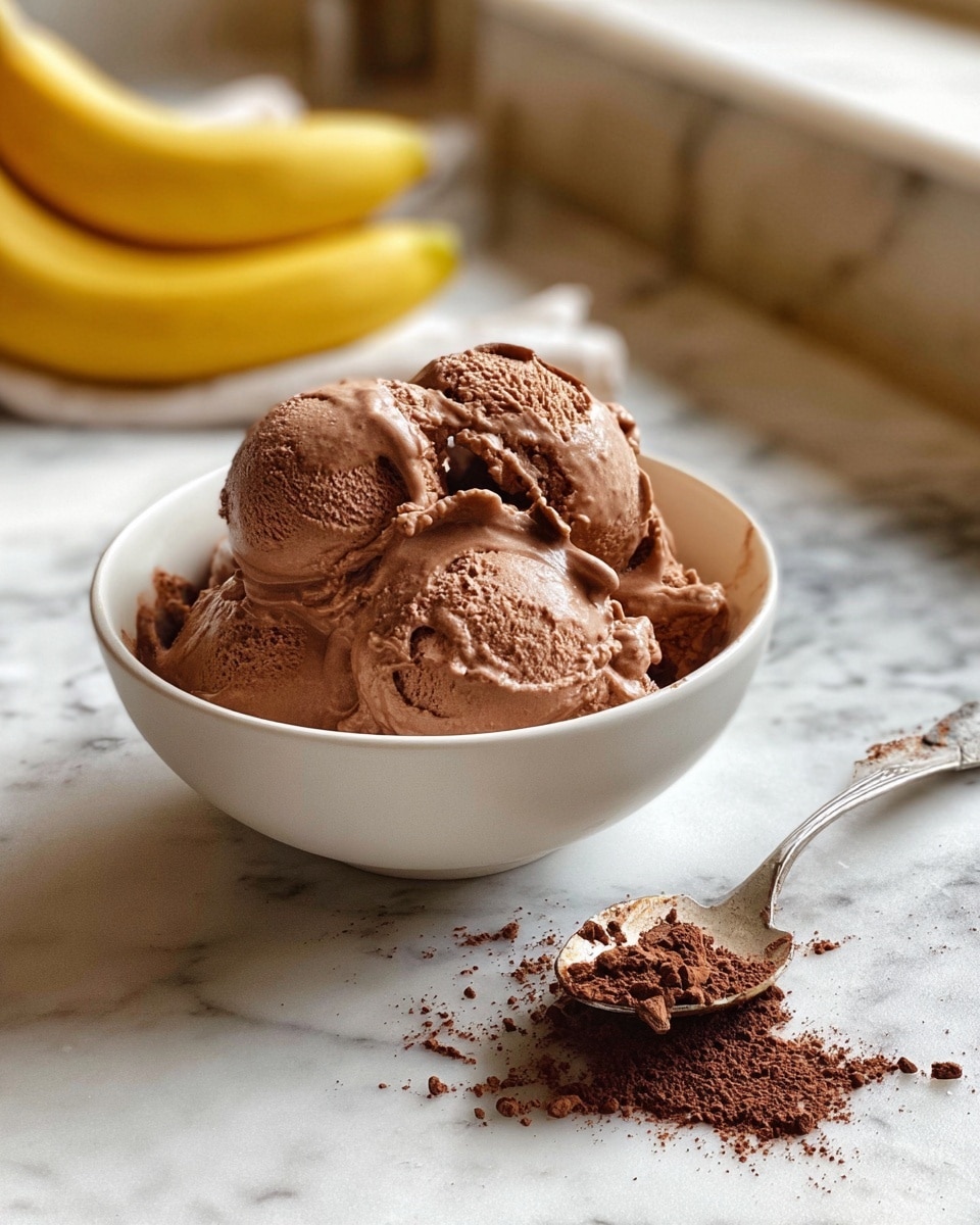 A white bowl filled with scoops of rich, creamy chocolate ice cream, piled loosely with smooth, soft texture and some melting at the edges, sits on a white marbled surface. Nearby, a silver spoon with traces of chocolate powder rests to the right, while a small pile of cacao powder is spread in front of the bowl. In the background, a bunch of bananas lies blurred, adding a soft yellow contrast. The scene shows a cozy, casual setting with natural lighting. Photo taken with an iphone --ar 4:5 --v 7
