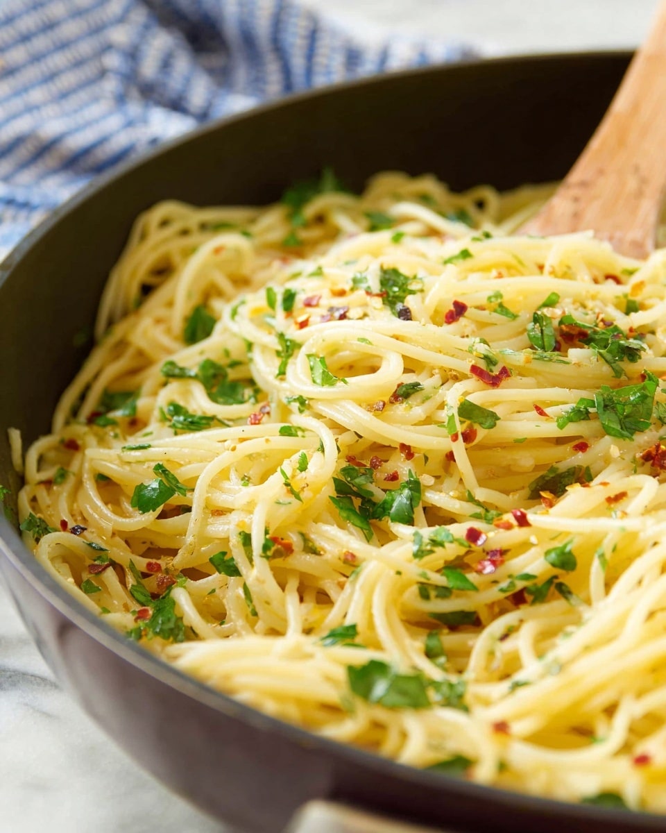 A close-up view of a pan filled with cooked spaghetti noodles, light yellow in color, mixed with scattered red chili flakes and finely chopped green parsley leaves spread evenly on top. The spaghetti strands are smooth and shiny, appearing slightly oily, with a wooden spoon visible in the pan stirring the noodles from the right side. The pan has a dark rim and is placed on a white marbled surface, with a blurred blue and white checkered cloth in the background. photo taken with an iphone --ar 4:5 --v 7