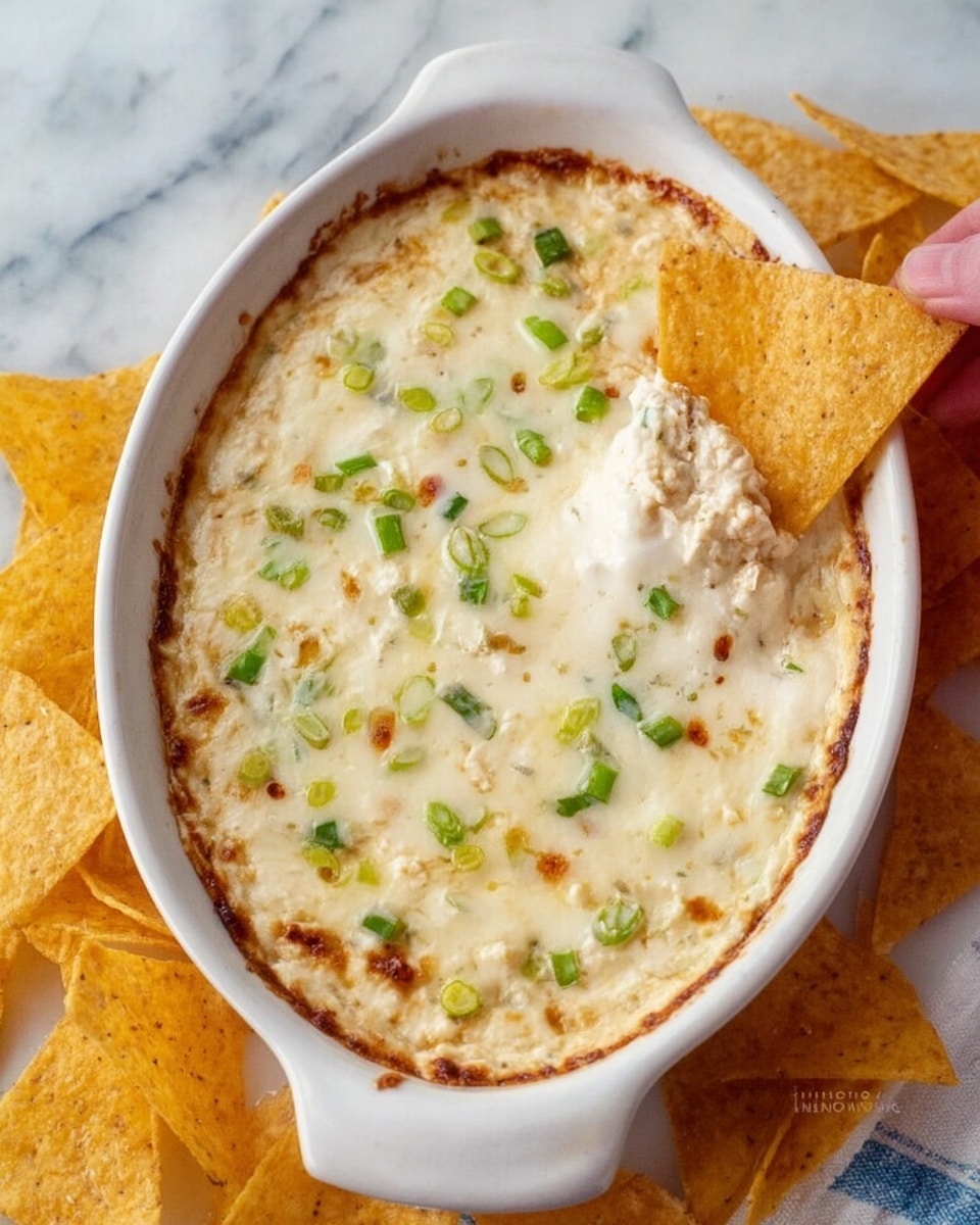 A white oval baking dish filled with a creamy, melted cheese dip that has a slightly browned edge. The top layer is smooth and glossy with small green onion pieces scattered evenly across the surface. Two triangular golden-brown chips are partially dipped on one side, showing the thick, creamy dip inside. The dish is placed on a white marbled surface, and a woman's hand is dipping one of the chips into the cheese. Additional golden chips surround the dish. Photo taken with an iphone --ar 4:5 --v 7