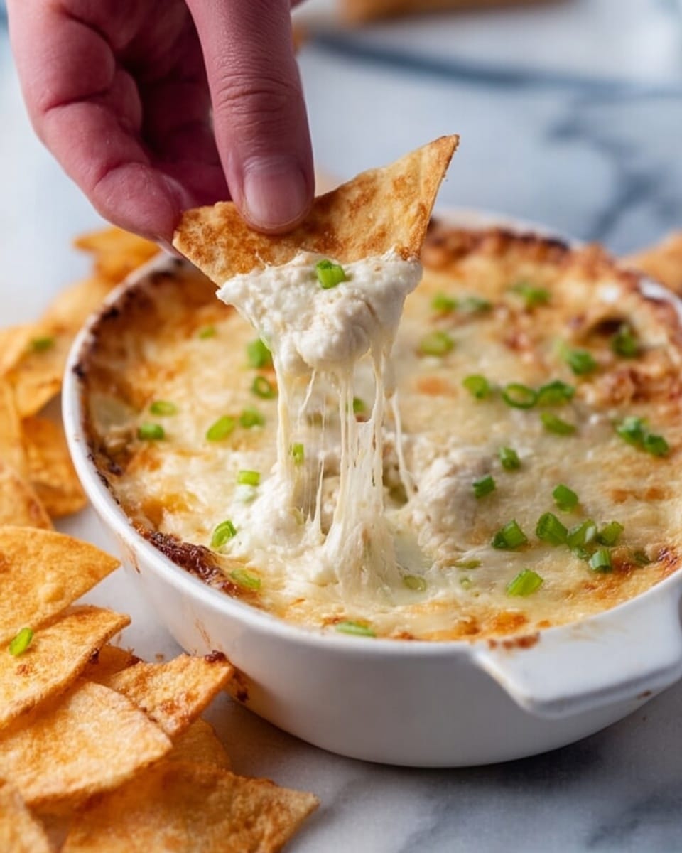 A close-up of a white oval dish filled with a creamy, melted cheese dip topped with small green onion pieces scattered across the surface. A woman's hand is lifting a triangular golden-brown chip dipped deep into the cheese, showing long, stretchy cheese strands pulling away from the dish. In the foreground, more golden chips with a crispy texture rest on a white marbled surface. Photo taken with an iphone --ar 4:5 --v 7