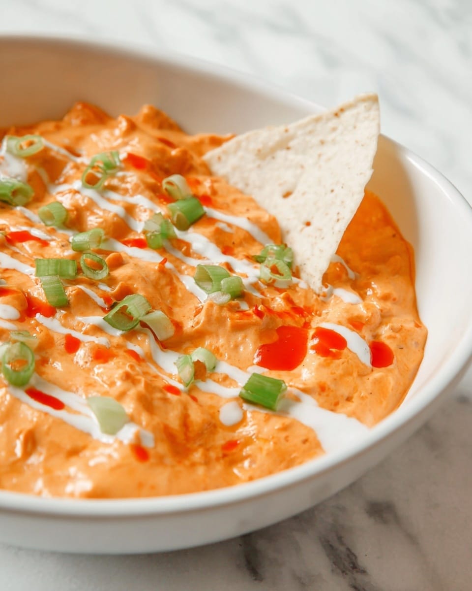 The image shows a creamy orange dip served in a white bowl. The dip has a thick and smooth texture with small chunks, topped with small green onion pieces and thin white sauce drizzles across the surface. There are also small drops of bright red sauce scattered on top. A white tortilla chip is partially dipped into the orange mixture, adding a crunchy element to the scene. The bowl sits on a white marbled surface. photo taken with an iphone --ar 4:5 --v 7