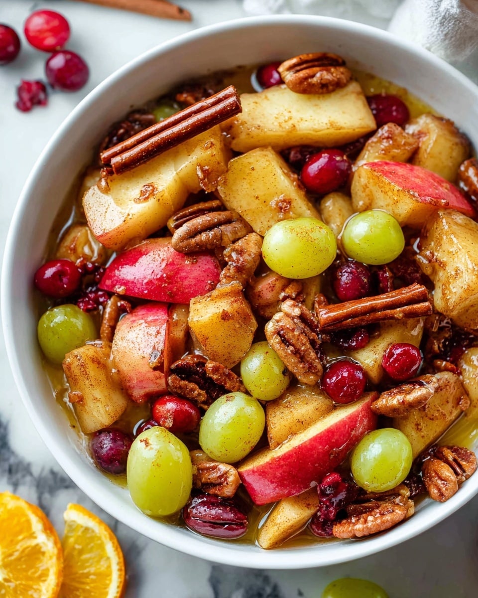 A close-up image of a white bowl filled with a warm fruit and nut mixture. The dish has many thick slices of cooked apple with red skin, slightly browned and soft. There are light brown slices of cooked pears mixed in, along with whole green grapes that have a smooth shiny surface. Bright red dried cranberries and several whole pecan halves are scattered throughout, adding texture. A few cinnamon sticks with a rough brown surface lie on top. The mixture is lightly coated with a glossy cinnamon-spiced liquid. The bowl rests on a white marbled textured surface with a partially visible slice of orange and green grapes to the side. Photo taken with an iphone --ar 4:5 --v 7