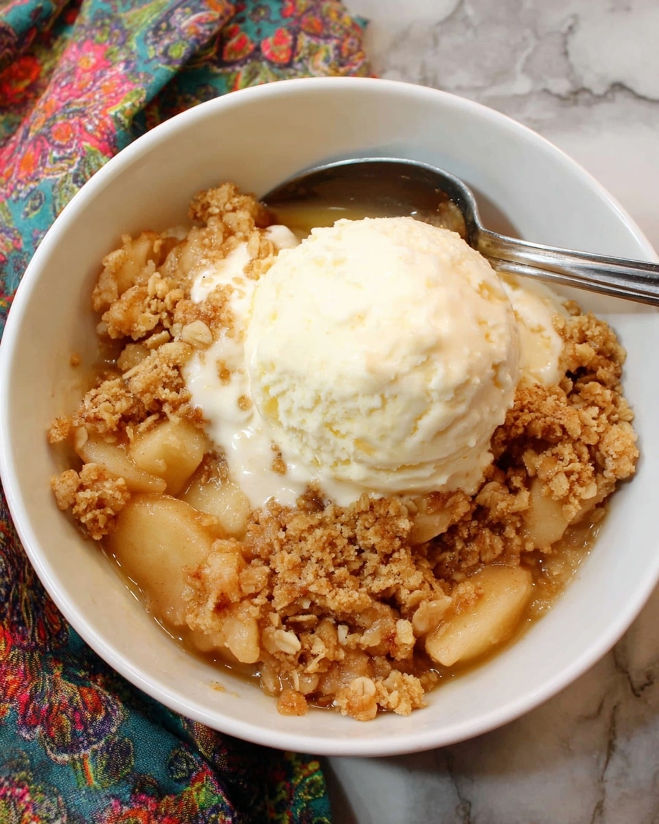 A white bowl filled with three visible layers: the bottom layer is soft, cooked apple pieces in a light golden sauce; the middle layer is a crumbly, golden-brown oat and sugar topping with a crunchy texture; the top layer is a large scoop of creamy, white vanilla ice cream that is beginning to melt slightly onto the warm crisp. A metal spoon rests inside the bowl, and the bowl sits on a surface with a white marbled texture and part of a colorful patterned cloth beside it. photo taken with an iphone --ar 4:5 --v 7
