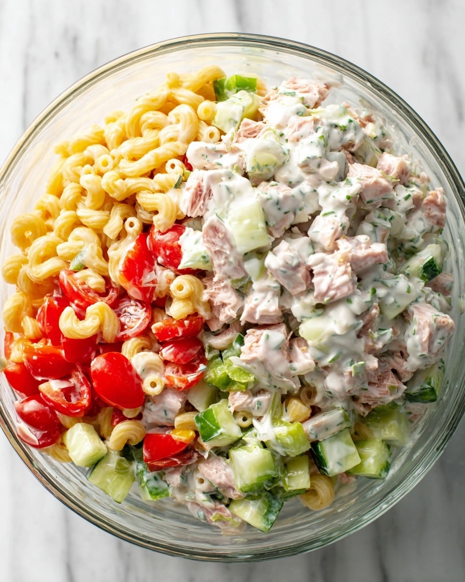 A close-up of a creamy pasta salad served on a white textured plate atop a white marbled surface. The dish has multiple layers: creamy white dressing coats small elbow macaroni pasta, chunks of light green cucumber, half slices of red cherry tomatoes, and small cubes of pale orange carrots, all mixed together. There is a sprinkle of black pepper and green herbs on top that add extra texture. A gold and white fork rests on the edge of the plate, partially under the salad. The photo taken with an iphone --ar 4:5 --v 7