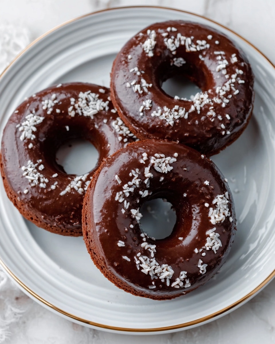 Three chocolate-glazed donuts sit on a white plate with a thin gold rim, placed on a white marbled surface. Each donut is covered evenly with smooth, shiny dark chocolate icing. White sprinkles are scattered on top of the chocolate layer, adding small light-colored spots. The donuts have a soft, slightly raised texture with a classic round shape and a hole in the center. Photo taken with an iphone --ar 4:5 --v 7