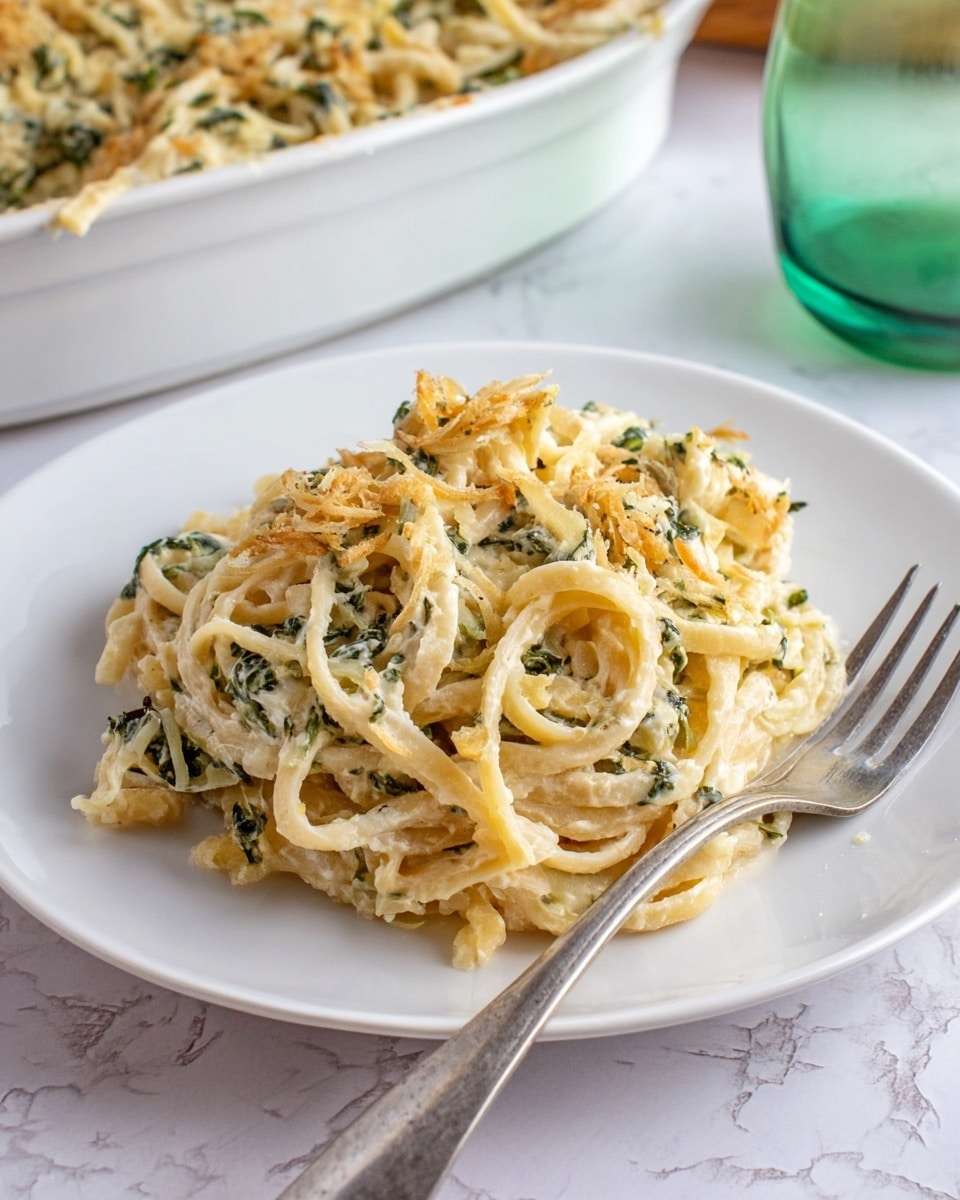 A white plate holds a pile of creamy pasta mixed with wilted green herbs and small crispy golden-brown bits scattered throughout. The pasta strands are thick and slightly twisted, coated in a light creamy sauce that adds a pale yellow tint to some pasta. A silver fork rests on the plate with its prongs pointing upward. In the background, a white casserole dish filled with more creamy pasta is partially visible on a white marbled surface, along with a green glass pitcher that reflects light softly. Photo taken with an iphone --ar 4:5 --v 7