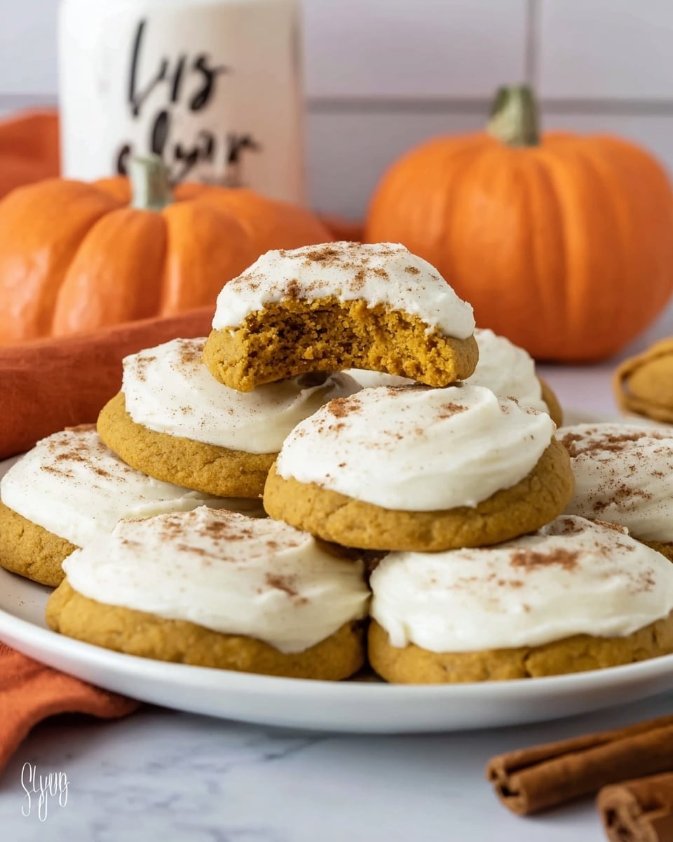 A white plate holds seven pumpkin cookies topped with a thick layer of creamy white frosting sprinkled with light brown cinnamon powder. One cookie is stacked on top of another in the center, with a bite taken out, showing the soft, orange-brown inside texture. Behind the plate are two small, bright orange pumpkins and a white sugar container with black lettering. The scene is set on a white marbled surface with an orange cloth and cinnamon sticks partially visible in the foreground. photo taken with an iphone --ar 4:5 --v 7