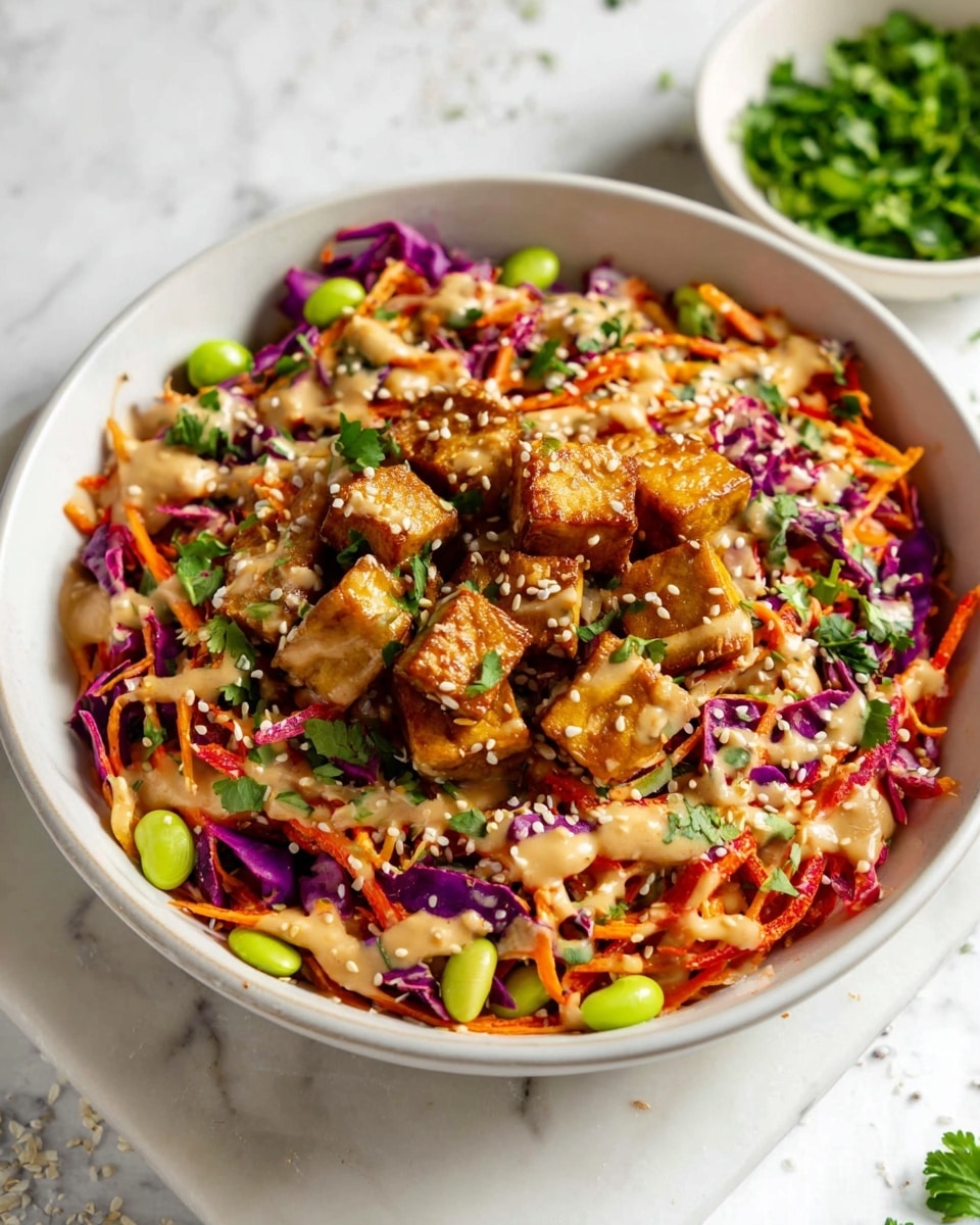 A white bowl filled with a colorful layered salad featuring pieces of golden-brown cooked tofu on top, mixed with thin strips of red and purple cabbage, orange carrot shreds, and light green edamame beans scattered around. The salad is drizzled with a creamy light brown sauce and sprinkled with chopped fresh cilantro and white sesame seeds, creating a textured and vibrant look. The bowl is set on a white marbled surface with a small white bowl of fresh green herbs visible in the background. Photo taken with an iphone --ar 4:5 --v 7