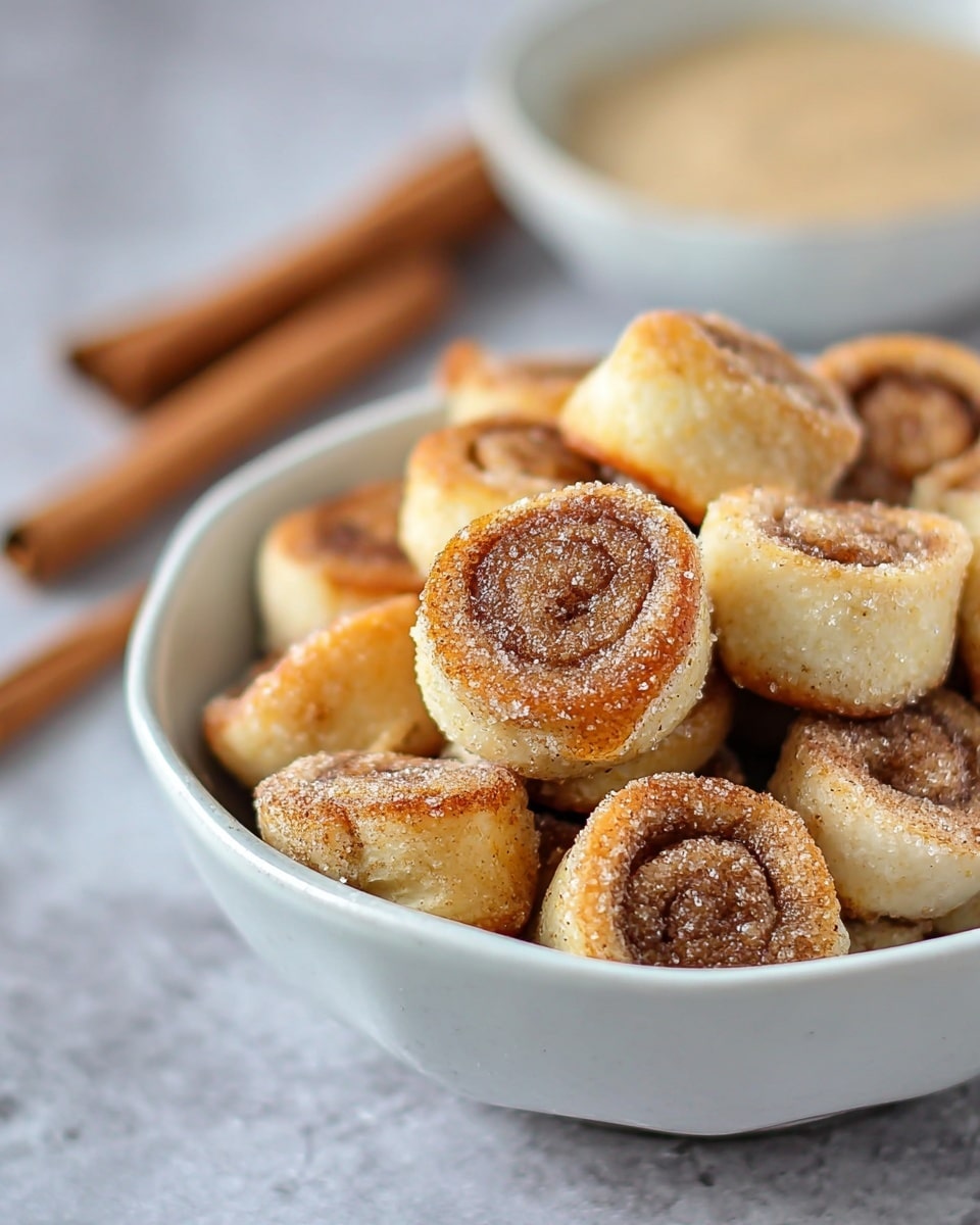 The image shows a white bowl filled with small cinnamon roll bites, each one having two visible layers: a light golden-brown outer dough and a darker cinnamon-sugar filling spiraled inside. The rolls appear soft with a slightly crispy edge, sprinkled lightly with sugar. In the blurred background, there is a white bowl containing a creamy beige sauce, and a couple of cinnamon sticks lay on the white marbled surface next to the bowl. The photo has a soft, natural light highlighting the texture of the rolls. photo taken with an iphone --ar 4:5 --v 7