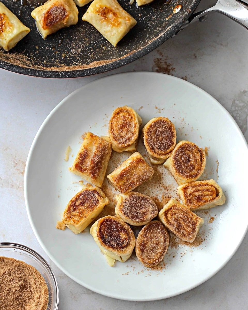 On a white plate with a white marbled background, there are about thirteen small rolled pastries cut into thick slices, each golden brown with a slightly crispy texture on the top, sprinkled with a light layer of cinnamon sugar giving a grainy, brown powdery look. Above the plate, a frying pan holds a few more of the rolled pastries cooking, showing a slightly shiny surface from butter or oil. In the lower corner, a clear bowl filled with cinnamon sugar is visible. Photo taken with an iphone --ar 4:5 --v 7