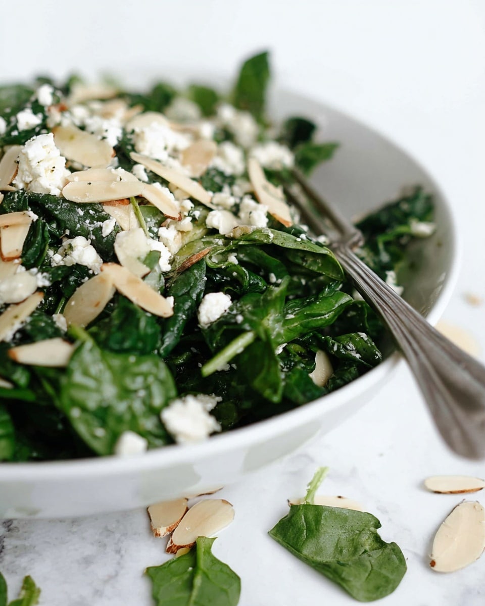 A white bowl filled with a fresh salad showing two layers: the bottom layer is dark green leafy spinach with a slightly glossy texture, and the top layer is scattered with small white crumbles of feta cheese, light beige almond slices, and thin green pea shoots. A silver fork rests inside the bowl, partially visible, and almond slices are scattered around the bowl on a white marbled texture. photo taken with an iphone --ar 4:5 --v 7