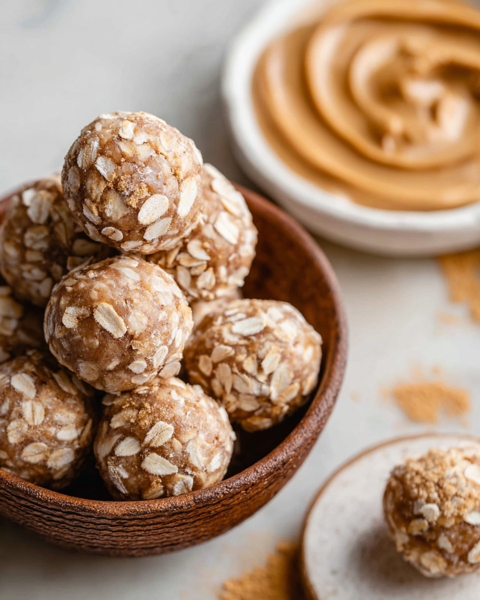 A close-up view of several round energy balls stacked inside a brown textured bowl, each ball covered in a layer of light beige oatmeal flakes with a dusting of cinnamon powder on top, showing a slightly rough texture. In the background, a blurred white bowl contains a swirl of creamy, light brown peanut butter, resting on a white marbled surface with faint golden-yellow streaks. At the bottom right, a white plate holds one of the oatmeal balls. photo taken with an iphone --ar 4:5 --v 7
