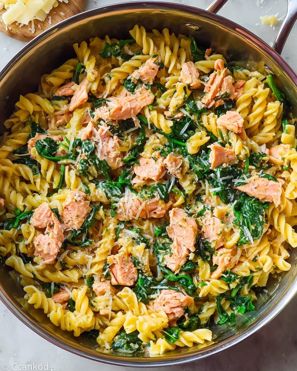 A close-up view of a pan filled with a pasta dish featuring three main layers: the base layer is yellow, twisted pasta with a wavy edge texture, mixed evenly throughout; the middle layer consists of bright green sautéed spinach leaves scattered across the pasta; the top layer includes chunks of pink cooked salmon placed randomly on top and mixed into the dish, with some grated white cheese sprinkled throughout. The pan is stainless steel and sits on a white marbled surface, with a few pieces of grated cheese around it. photo taken with an iphone --ar 4:5 --v 7