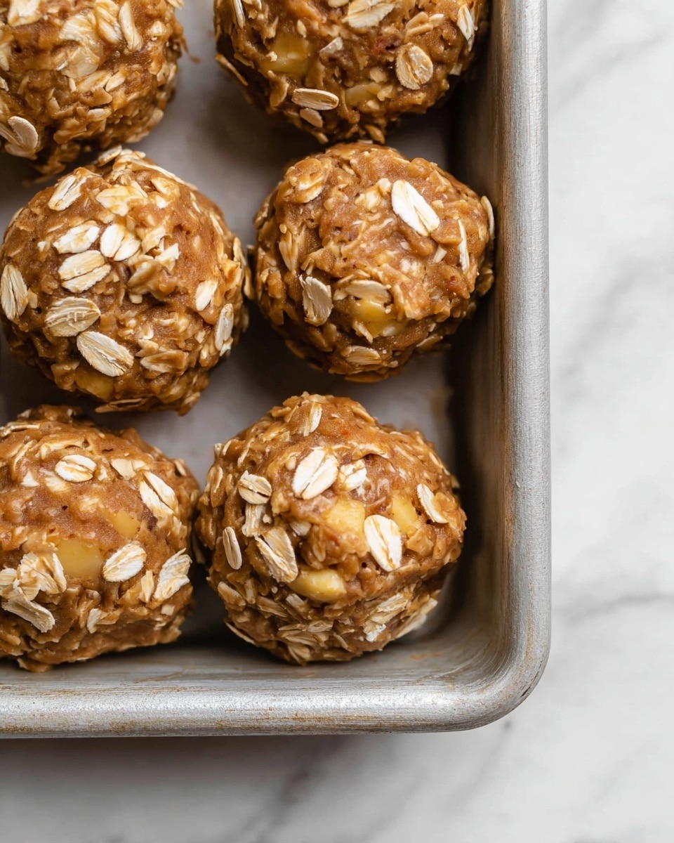 The image shows several round cookie dough balls placed closely together in a metal baking tray on a white marbled surface. Each dough ball is thick and chunky, with a soft brown base layer mixed with visible chunks of light yellow apple pieces embedded throughout. Rolled oats are sprinkled on top and slightly pressed into the dough, adding a textural contrast with their flaky, off-white color. The dough appears moist and dense, with a slightly rough texture. The baking tray is simple and metallic, contrasting with the natural tones of the dough balls. photo taken with an iphone --ar 4:5 --v 7