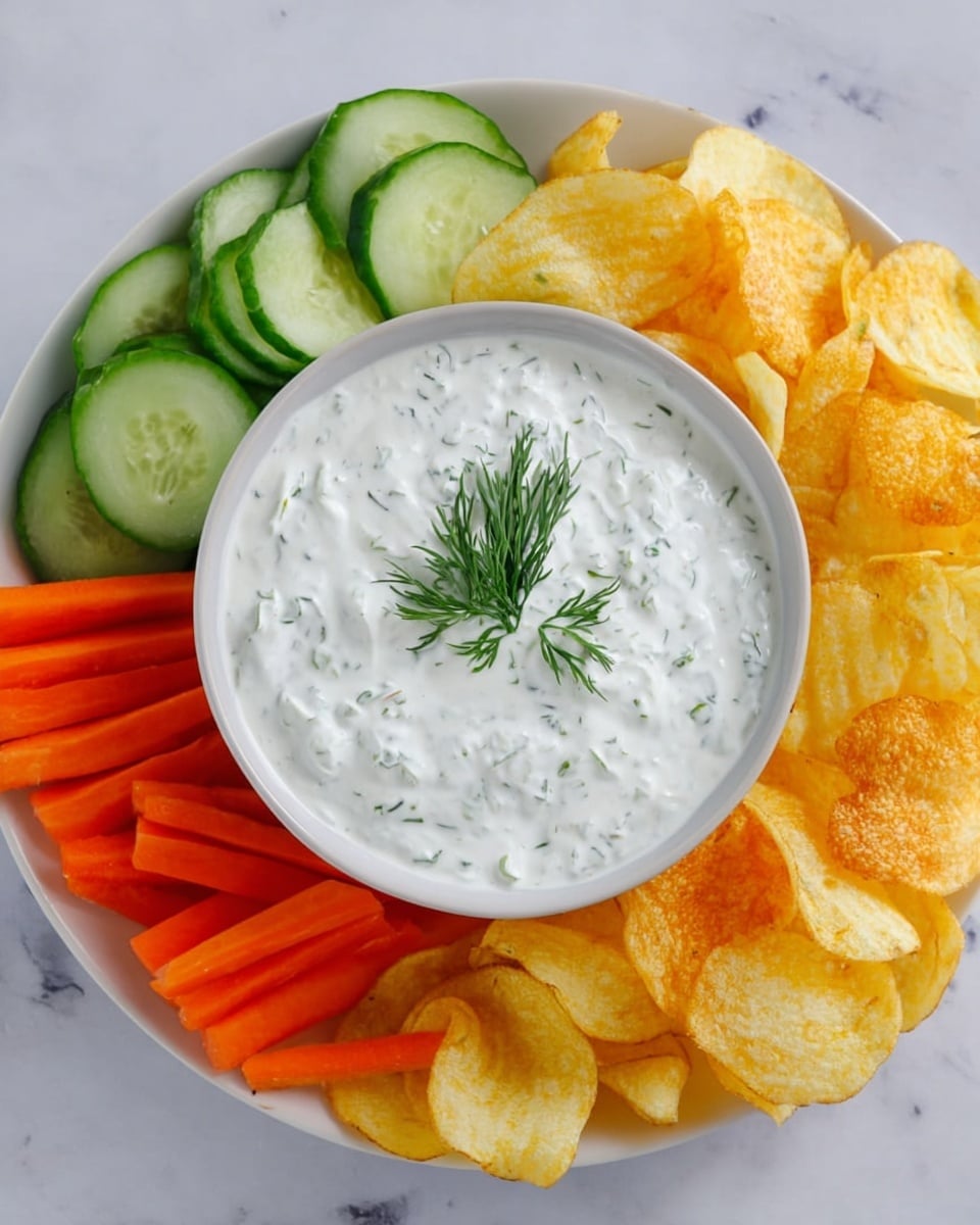 A white plate on a white marbled surface holds a small bowl at the center filled with creamy white dip speckled with green herbs, topped with a small sprig of fresh dill. Surrounding the bowl are fresh cucumber slices with dark green edges and light green centers, bright orange carrot sticks arranged in a small pile, and golden-yellow potato chips with a crispy texture. The layers create a colorful and fresh snack presentation. photo taken with an iphone --ar 4:5 --v 7