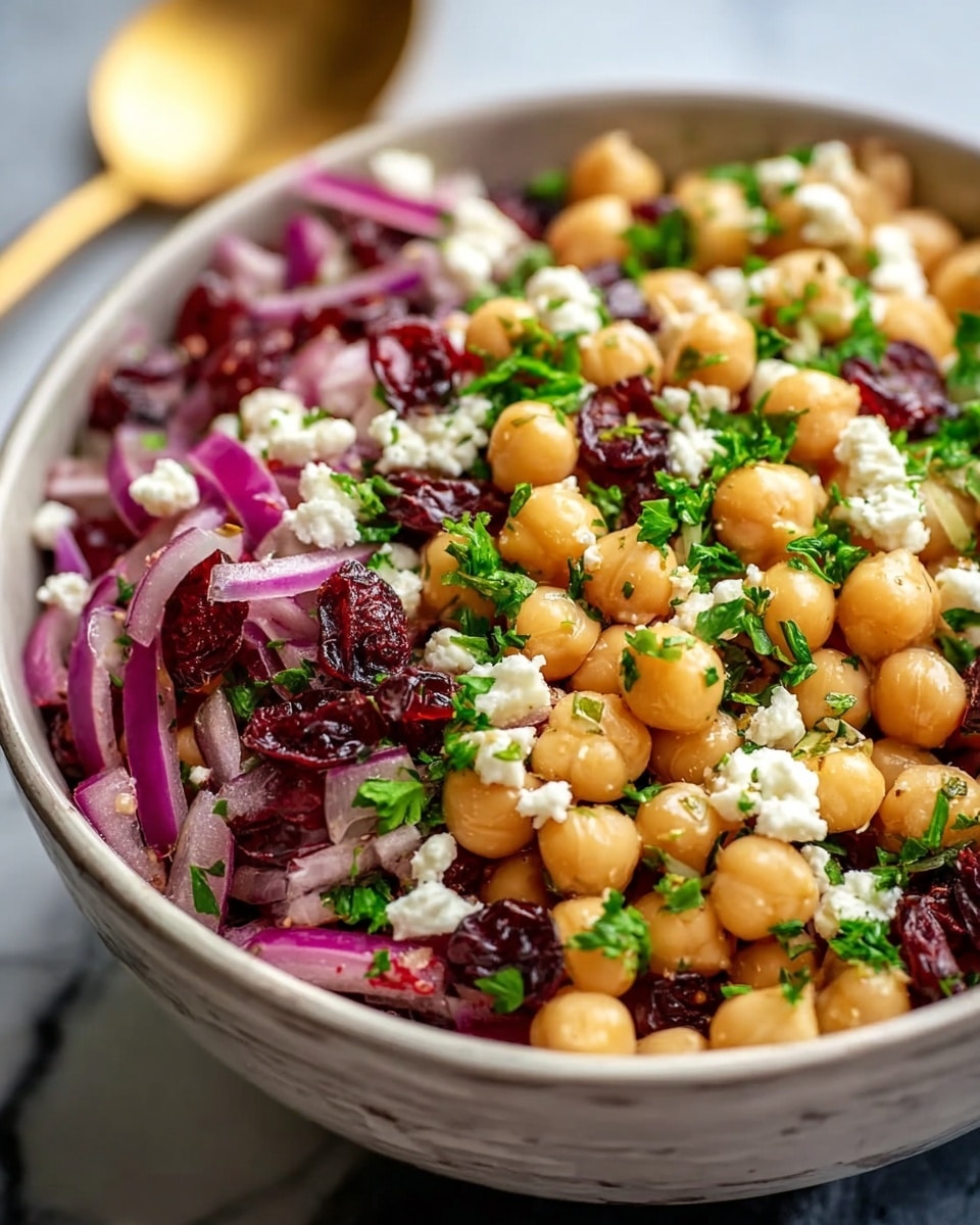 A close-up view of a salad served in a white bowl filled with chickpeas forming the main layer, soft beige in color and round in shape. Mixed throughout are bright red dried cranberries and crumbled white cheese sprinkled evenly over the top. Thin slices of purple onion are placed on the surface, adding contrast along with green chopped herbs scattered all over the dish, giving it a fresh and colorful look. The background shows a white marbled texture. Photo taken with an iphone --ar 4:5 --v 7
