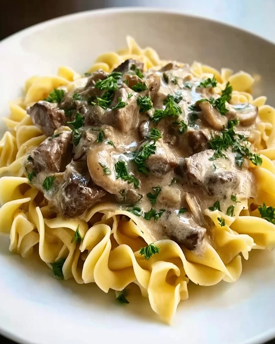 The image shows a white plate filled with yellow curly pasta at the bottom. On top of the pasta, there is a thick layer of creamy brown sauce with chunks of dark brown mushrooms mixed in. The sauce looks rich and smooth, covering the pasta well. Small green parsley leaves are sprinkled on top, adding a fresh touch of color. The plate sits on a white marbled surface. Photo taken with an iphone --ar 4:5 --v 7
