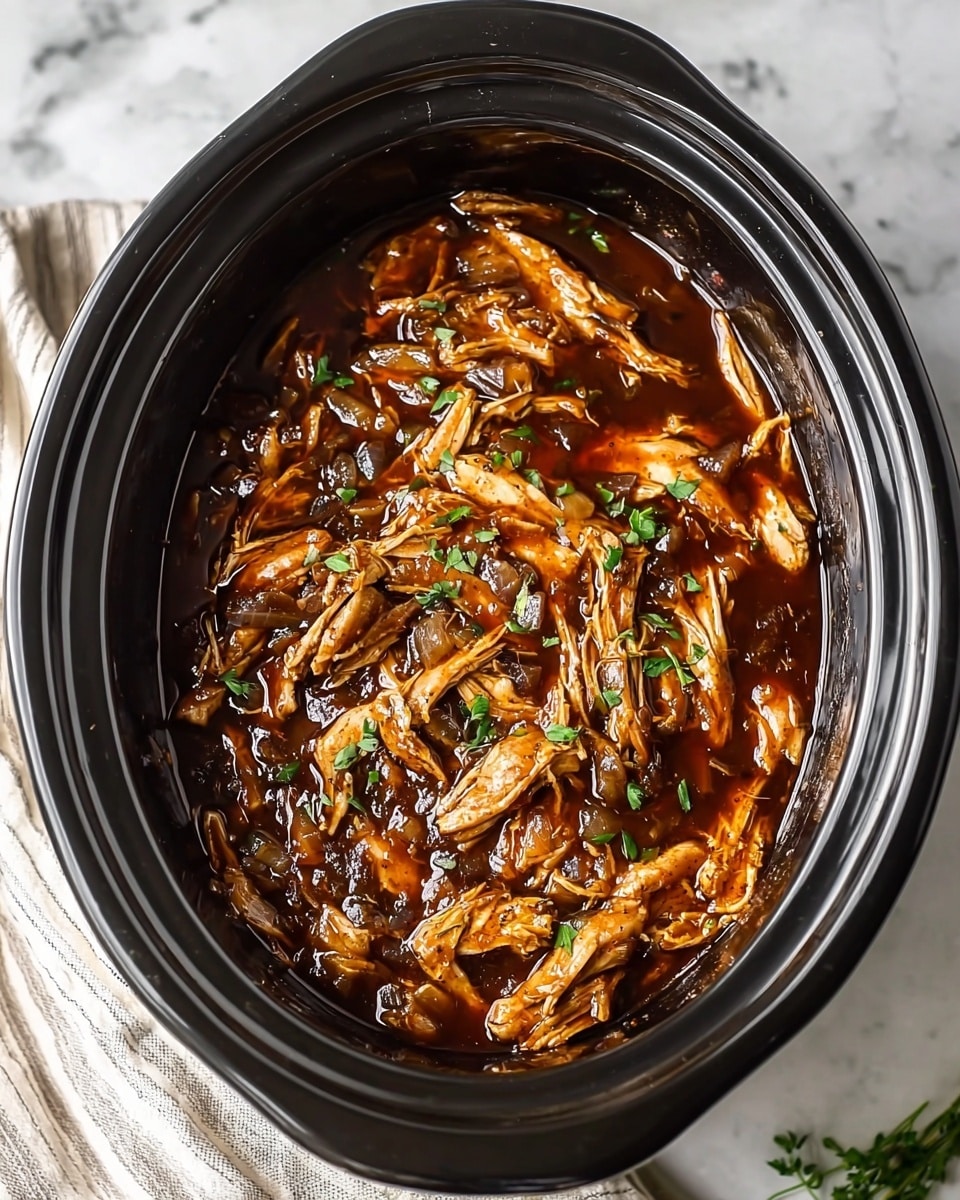 A black slow cooker filled with shredded chicken covered in a thick, glossy dark brown sauce. The chicken pieces are layered unevenly with small bits of cooked onions mixed throughout, and the dish is topped with small, bright green chopped herbs for color contrast. The slow cooker sits on a white marbled surface with a soft striped cloth partially visible on one side. photo taken with an iphone --ar 4:5 --v 7