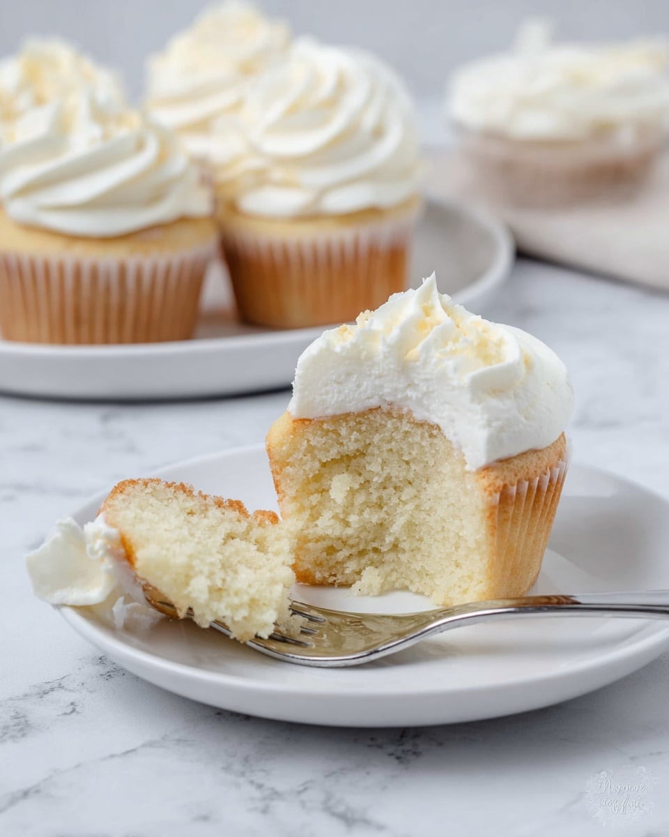 A group of vanilla cupcakes is placed on a white plate over a white marbled surface. Each cupcake has a light golden base with a soft texture, topped with a single fluffy swirl of smooth white frosting that has a slightly glossy finish. The frosting is piped in a spiral shape starting from the outer edge and ending in the middle, creating a rose-like pattern on top. The cupcakes are arranged close to each other, showing the detail on the frosting swirl and the gentle crumb of the cupcake beneath. Photo taken with an iphone --ar 4:5 --v 7