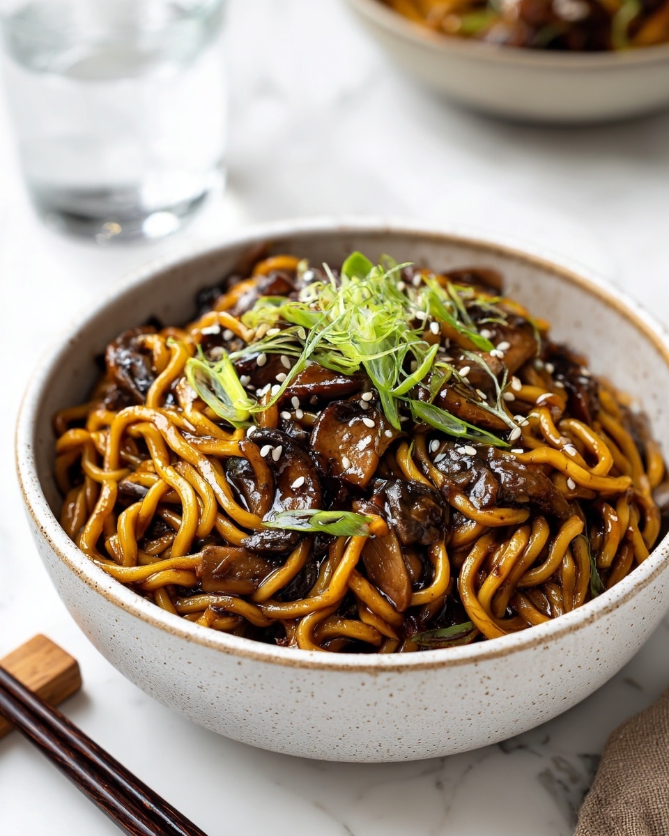 A white speckled bowl filled with thick, twisted golden-brown noodles coated in a glossy dark sauce, mixed with chunks of tender brown mushroom pieces. The noodles are topped with thinly sliced bright green scallions and sprinkled with small white sesame seeds, adding contrast. The bowl is placed on a white marbled surface with a pair of dark chopsticks resting on a light wood chopstick holder nearby. A glass of water and another similar bowl are blurred in the background. photo taken with an iphone --ar 4:5 --v 7