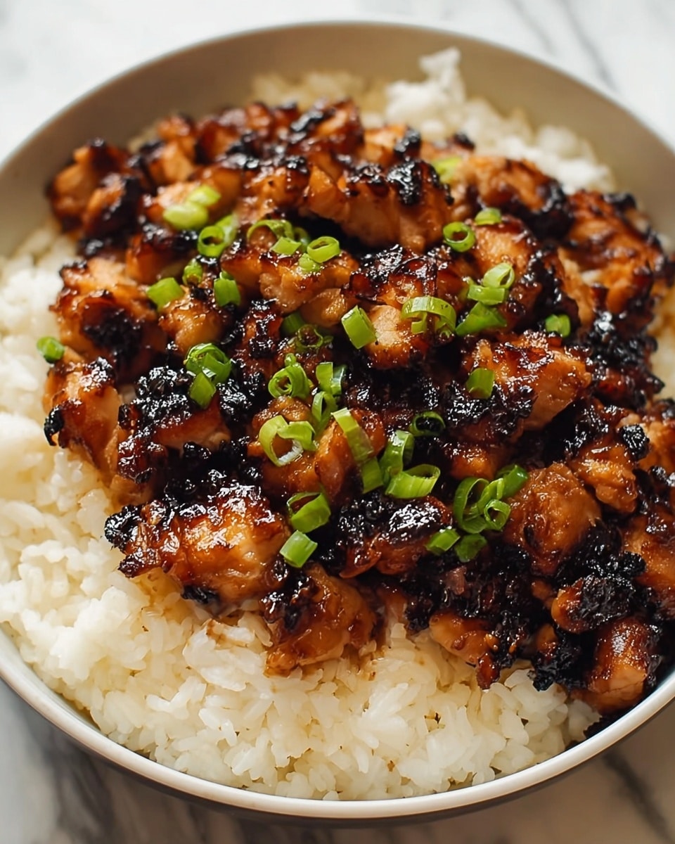 A close-up of a bowl filled with two layers: the bottom layer is fluffy white rice with a soft texture, while the top layer is made up of small dark brown grilled chicken pieces coated in a shiny glaze, showing some charred black marks. The chicken is garnished with small chopped green onions scattered on top. The bowl itself is white, sitting on a white marbled surface. photo taken with an iphone --ar 4:5 --v 7