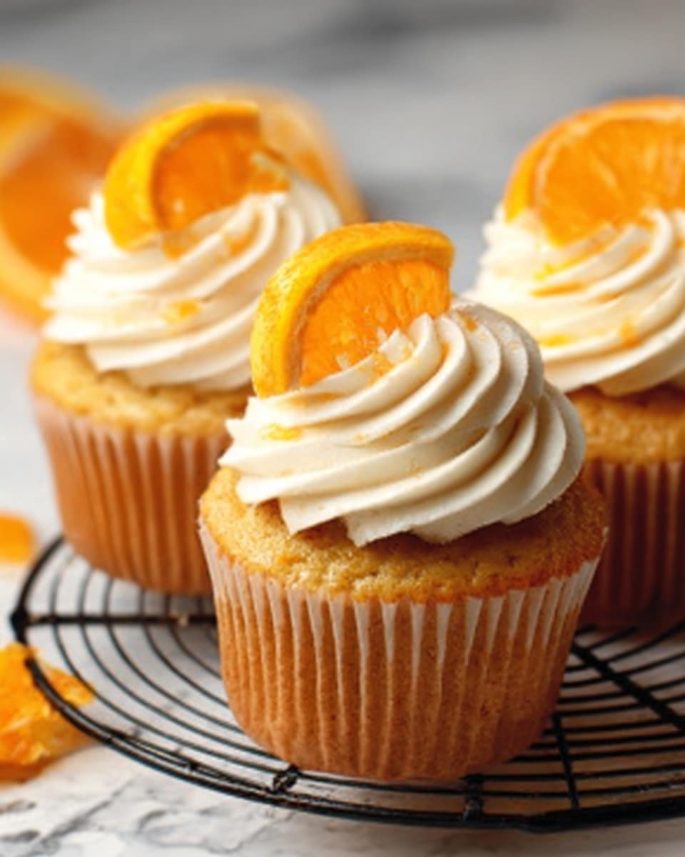 Three orange cupcakes sit on a round black wire rack on a white marbled surface. Each cupcake has a light brown base topped with a swirl of white frosting. On top of each frosting swirl rests a small orange slice, adding a bright pop of color. The texture of the frosting looks smooth and fluffy while the cupcake base appears moist and soft. Photo taken with an iphone --ar 4:5 --v 7
