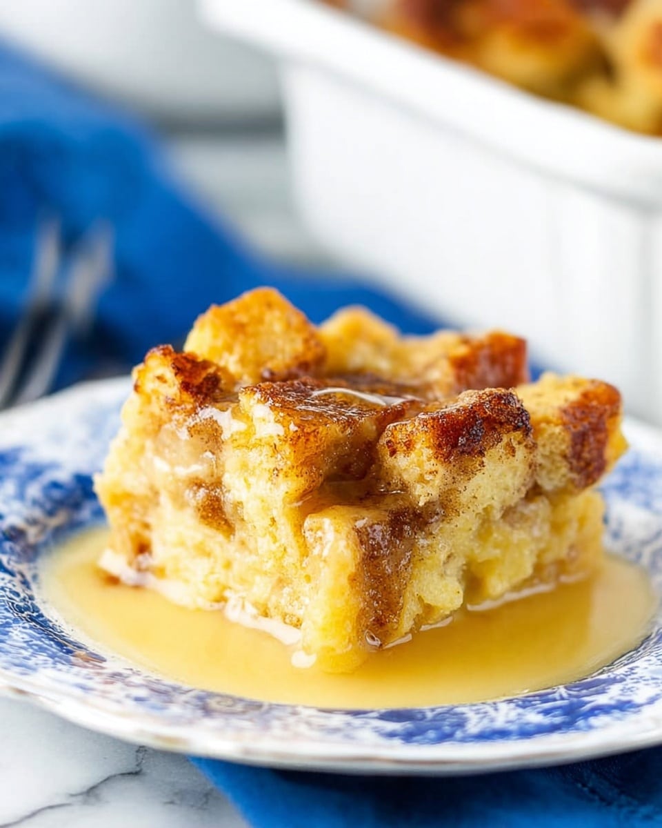 A close-up single square piece of bread pudding on a white decorative plate with a blue cloth underneath. The bread pudding has one main visible layer made of soft, golden-yellow, slightly chunky baked bread soaked in custard with a light brown cinnamon swirl through the middle. The top layer shows slightly crispy browned edges and small soft bread cubes that rise above the base layer. A thick, shiny, pale yellow sauce is pooling around the bread pudding piece on the plate. The background shows a blurred white marbled surface and a white dish of more bread pudding behind it. Photo taken with an iphone --ar 4:5 --v 7