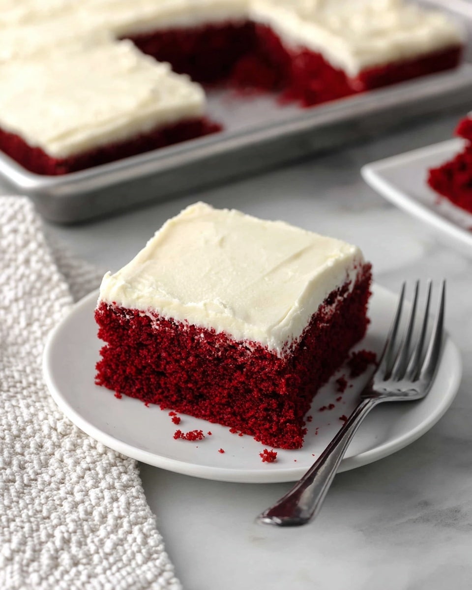 A square slice of red velvet cake sits on a white plate, showing two clear layers— a thick, deep red soft cake base and a smooth, creamy white frosting layer on top. The cake slice is moist with a slightly crumbly texture, while the frosting appears thick and evenly spread. In the background, a metal baking tray with more cake is visible, with one piece missing. The scene is set on a white marbled surface with a silver fork nearby and a textured white cloth partially visible on the left side. photo taken with an iphone --ar 4:5 --v 7