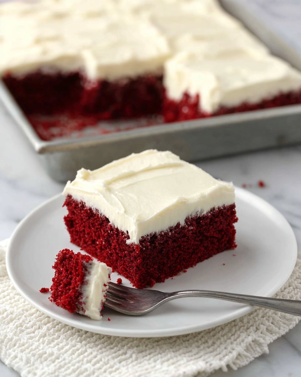 A single square piece of red velvet cake sits on a white plate, showing two clear layers: a thick, moist, dark red sponge base and a smooth, creamy white frosting spread evenly on top. In the foreground, a silver fork holds a smaller bite-sized piece of the cake with both the red sponge and white frosting visible. Behind the plate, the larger cake rests in a metal baking pan with a missing piece, revealing the same red and white layers. All elements are placed on a white marbled surface with a textured white cloth nearby. Photo taken with an iphone --ar 4:5 --v 7