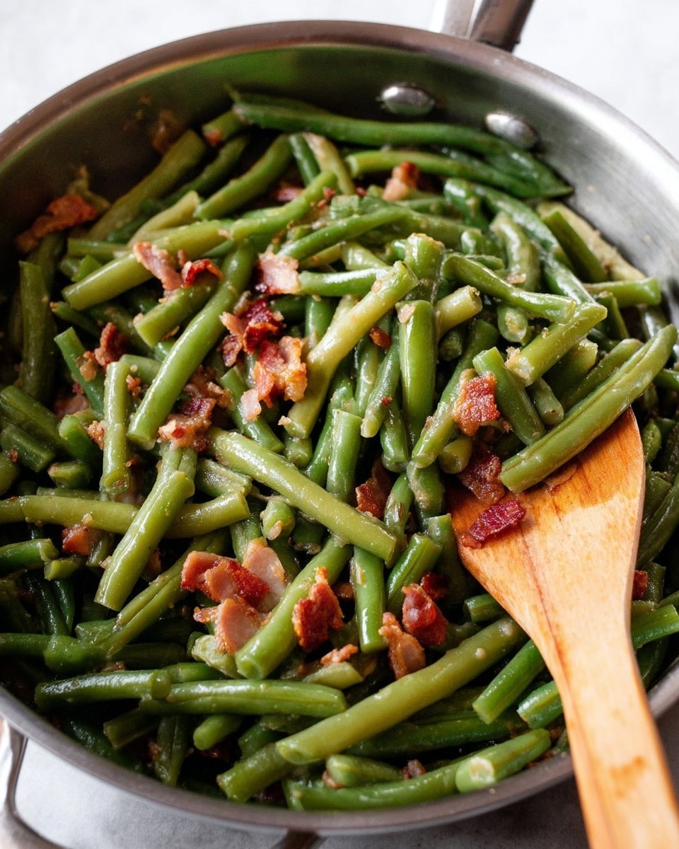 The image shows cooked green beans mixed with small pieces of reddish-brown bacon inside a silver metal pan. The green beans are cut into medium-sized pieces and are a deep, soft green color with a shiny, moist texture. The bacon bits are scattered unevenly throughout the green beans, adding contrast with their crisp, slightly oily look. A light brown wooden spoon rests inside the pan on the right side, partially covering some green beans. The pan's metal surface is smooth and reflective. The scene is set on a white marbled surface. photo taken with an iphone --ar 4:5 --v 7