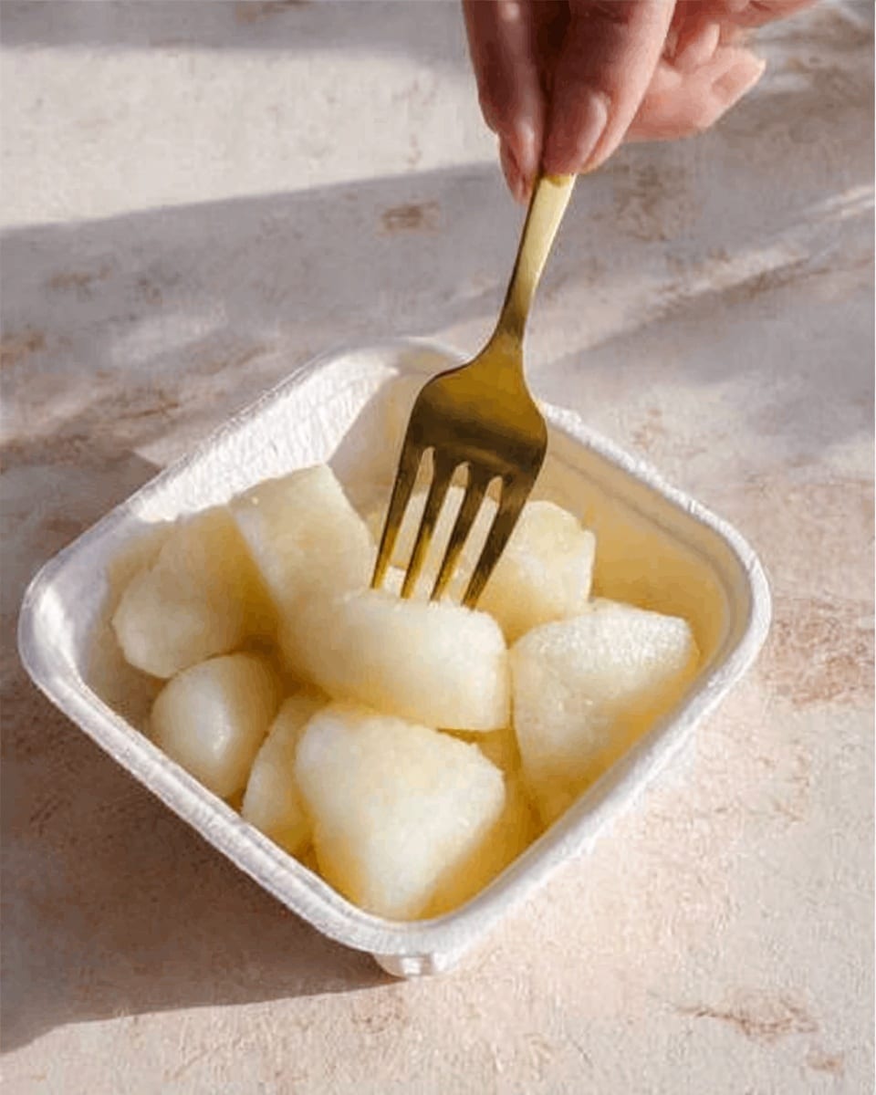 The image shows a white foam container filled with several pieces of peeled white fruit or rice cakes, arranged closely together. A golden fork is picking up one piece, held by a woman's hand from the top. The background surface is a white marbled texture with soft natural light giving a warm tone to the scene. Photo taken with an iphone --ar 4:5 --v 7