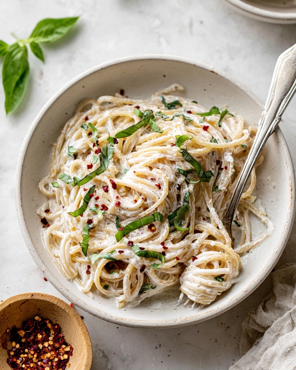 A white bowl filled with creamy spaghetti coated in a white sauce, sprinkled with thin strips of fresh green basil and a few red pepper flakes. The noodles are twisted around a silver fork resting inside the bowl, showing layers of smooth, glossy sauce covering the pasta. The bowl sits on a white marbled surface, accompanied by a small light wooden bowl with more red pepper flakes nearby and a few green basil leaves in the top left corner. photo taken with an iphone --ar 4:5 --v 7