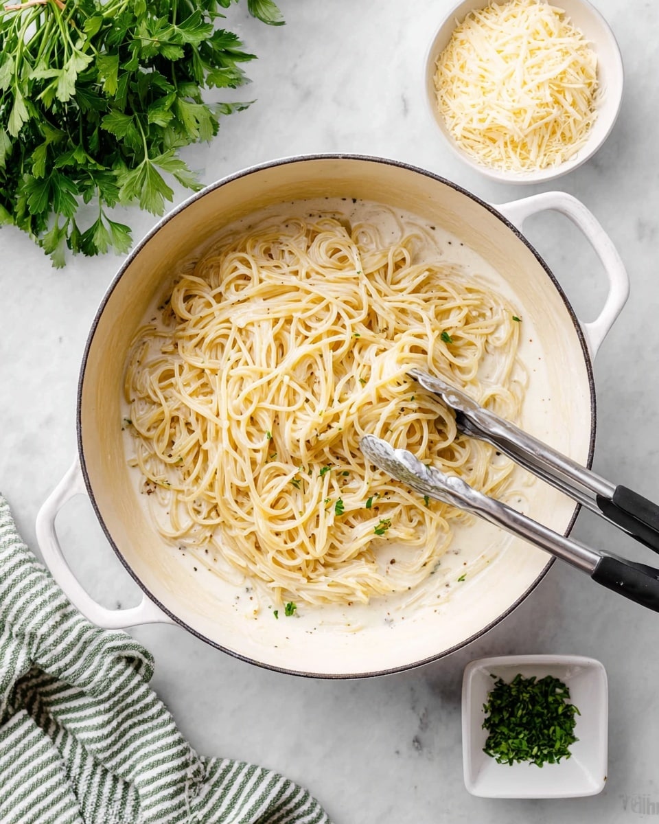 A white pot filled with creamy white sauce and long, light yellow pasta strands is centered on a white marbled surface. The pasta is lifted slightly by silver tongs with black handles, creating a small mound in the middle. To the top right, there is a small white bowl with grated pale yellow cheese, and below it a tiny square white dish holding finely chopped green herbs. On the left side, part of a green-and-white striped cloth lies next to fresh green parsley. Photo taken with an iphone --ar 4:5 --v 7