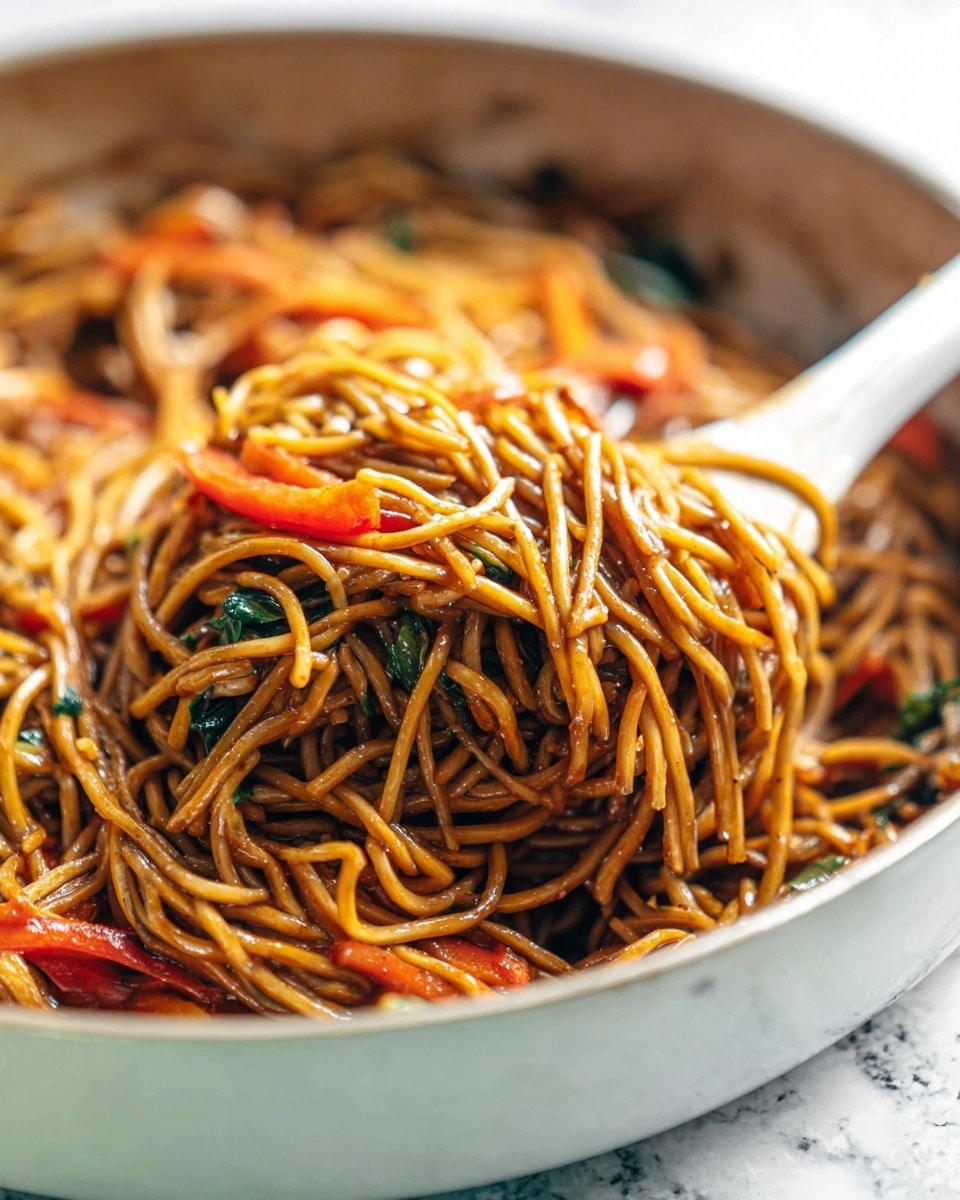 A close-up of a large pile of cooked noodles with a glossy brown sauce, mixed with thin slices of red bell pepper and small bits of green leafy vegetables, all resting inside a white skillet with a visible white spatula lifting some noodles, the noodles look soft and tangled with a slightly oily sheen, on a white marbled texture surface. photo taken with an iphone --ar 4:5 --v 7