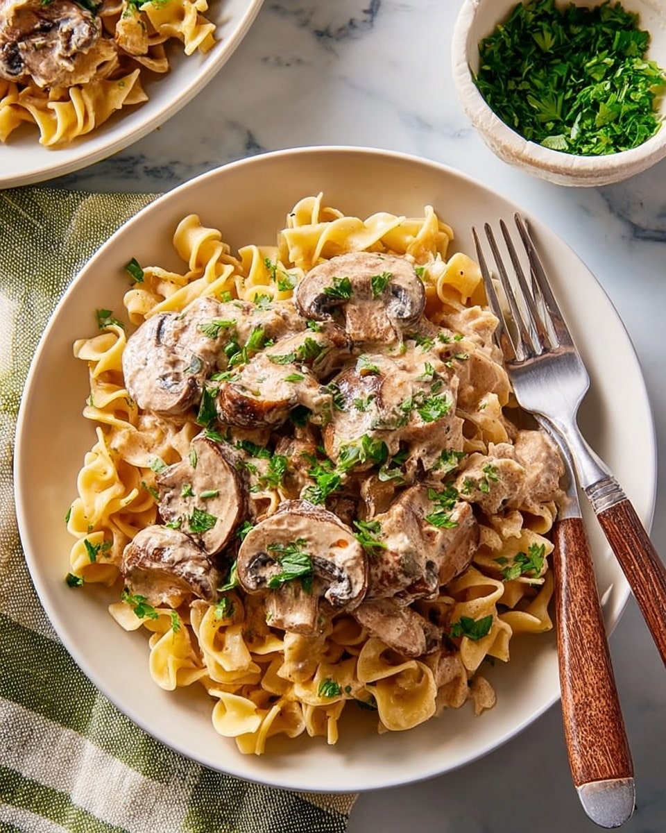 A dark plate holds a dish with three main layers: the base is a bed of pale yellow twisted egg noodles, topped with a creamy beige sauce mixed with slices of light brown cooked mushrooms, and garnished with small green parsley leaves scattered on top. A fork with a wooden handle rests on the right side of the plate. The plate sits on a white marbled surface next to a striped cloth napkin, with another plate of the same dish and a small bowl of chopped green parsley nearby. photo taken with an iphone --ar 4:5 --v 7