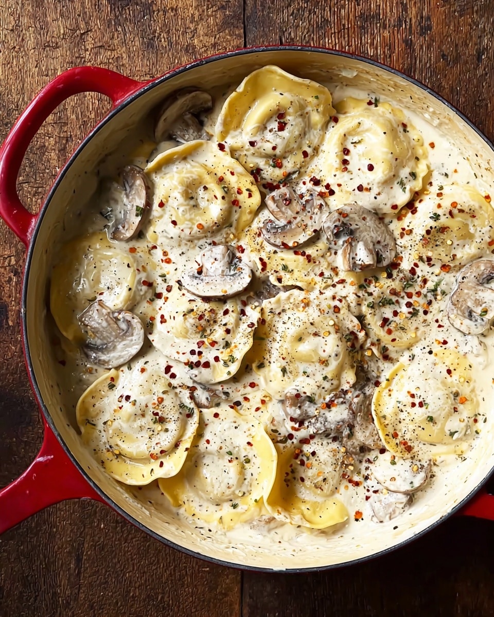 A round red pot filled with large, soft rounds of yellow ravioli pasta covered in a thick white creamy sauce with visible slices of light brown mushrooms scattered on top. The sauce is speckled with black pepper and red chili flakes, giving a textured look across the dish. The pasta is gently layered and partly submerged in the sauce, with some folds visible on the edges. The pot sits on a textured brown wooden surface. photo taken with an iphone --ar 4:5 --v 7