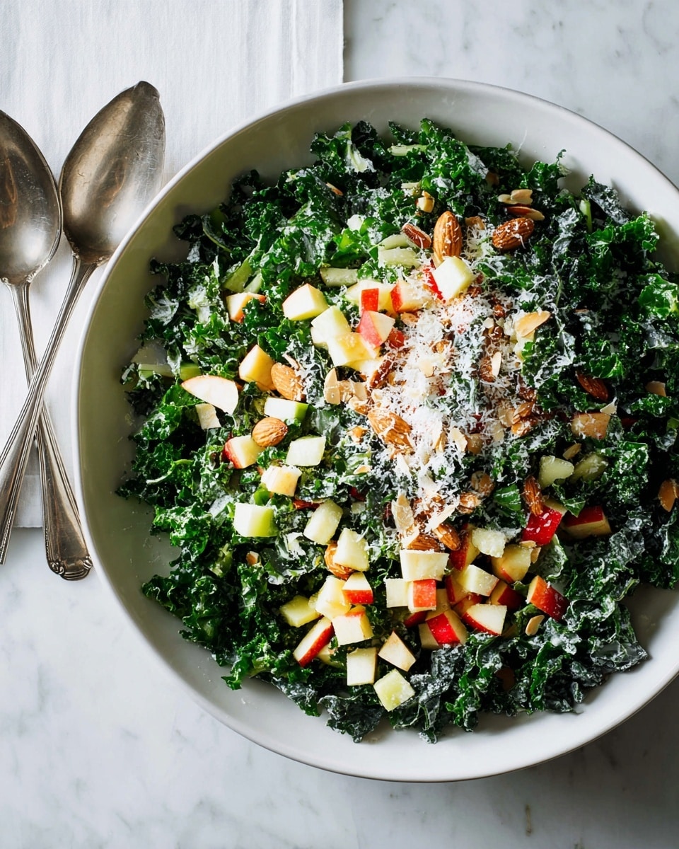 A large white bowl filled with a fresh kale salad, showing three main layers: the base layer of dark green, curly kale leaves spread evenly, a middle layer of small cubed apple pieces in red and pale yellow tones, mixed with chopped light brown almonds, and a top layer sprinkled with finely grated white cheese, creating a soft texture over the salad; the bowl is placed on a white marbled surface with two silver salad serving spoons resting nearby. photo taken with an iphone --ar 4:5 --v 7