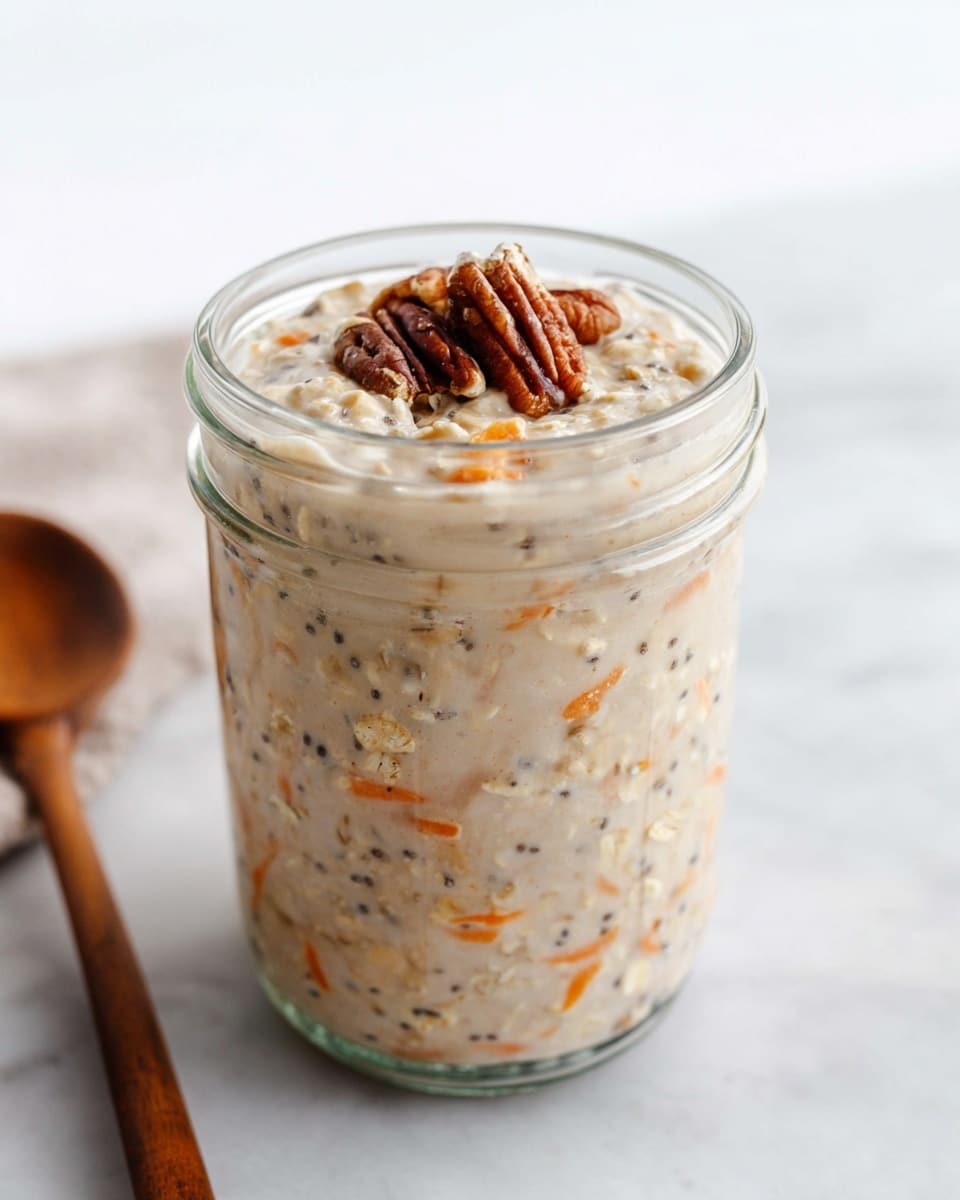 A clear glass jar filled with creamy oatmeal showing soft, mixed textures of light beige with small orange carrot bits and tiny dark chia seeds evenly spread throughout. The oatmeal reaches near the top of the jar, and a few small pecan pieces with a rich brown color sit on top as garnish. The jar is placed on a white marbled surface with a wooden spoon blurred in the background, creating a clean and simple look. Photo taken with an iphone --ar 4:5 --v 7